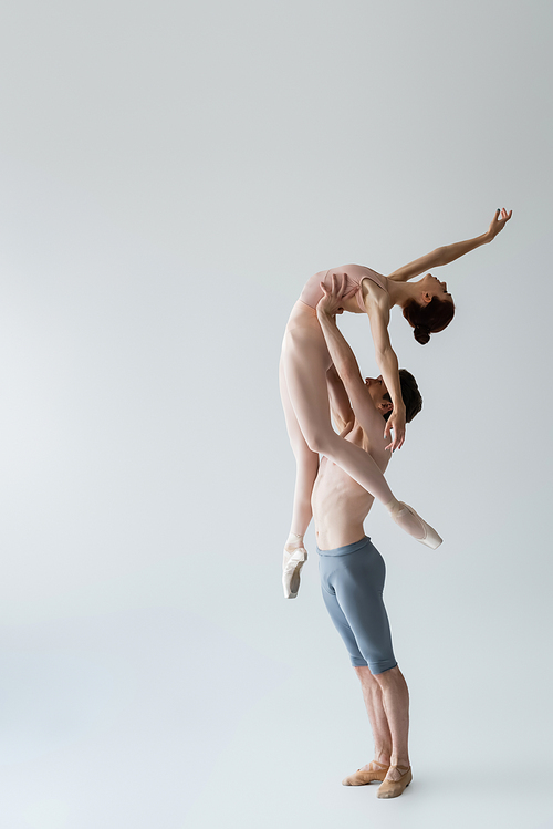 full length of shirtless ballet dancer lifting young ballerina on grey