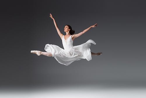 full length of graceful woman in white dress jumping on dark grey