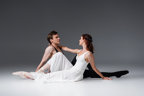 full length of graceful ballerina in white dress sitting with smiling partner on dark grey