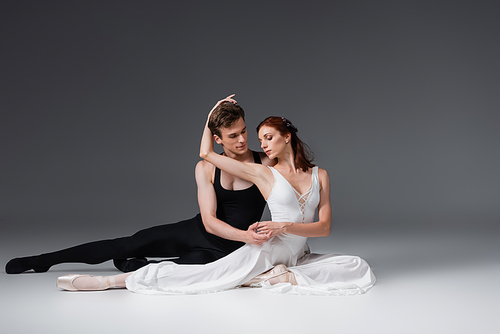 full length of flexible dancer and graceful ballerina in white dress sitting on dark grey