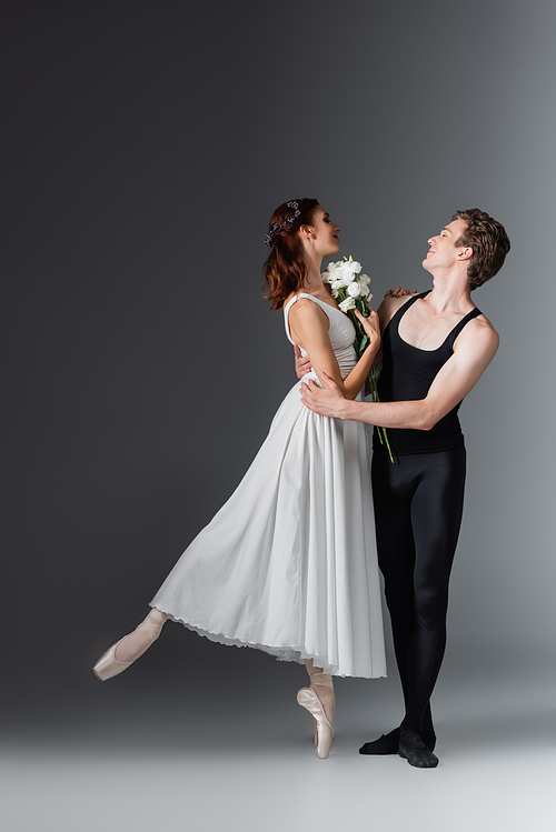 full length of graceful ballerina in white dress holding flowers and dancing with partner on dark grey