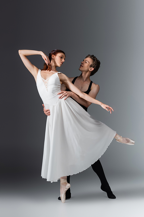 full length of young ballerina in white dress dancing with partner on dark grey