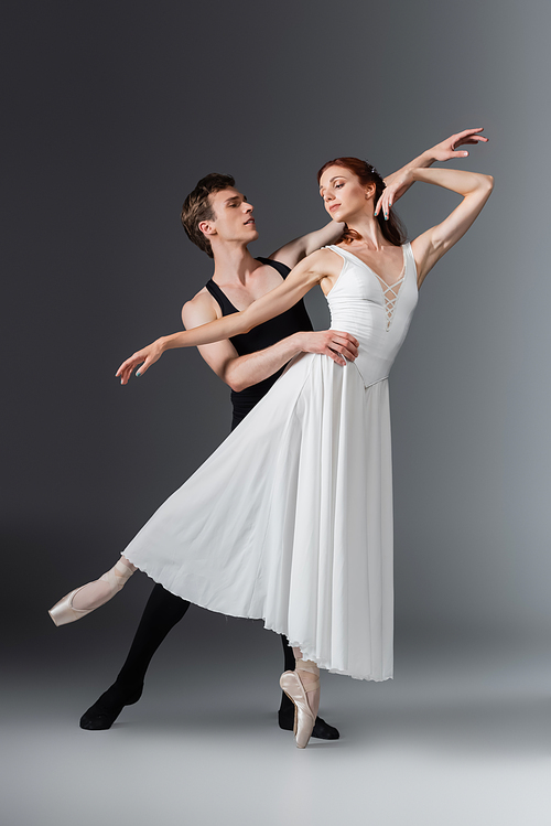 full length of young graceful ballerina in white dress dancing with partner on dark grey