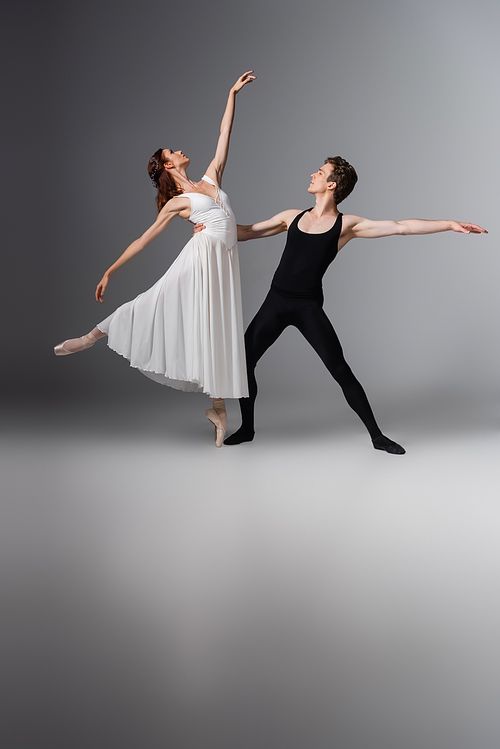 full length of graceful ballerina in white dress dancing with partner on dark grey