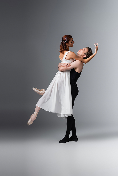 full length of dancer lifting graceful ballerina in white dress on dark grey
