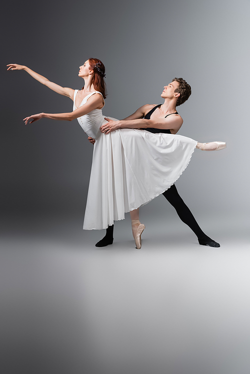 full length of graceful ballerina in white dress dancing with young partner on dark grey