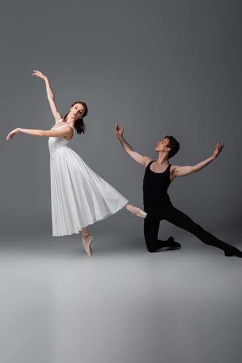 full length of graceful ballerina in white dress dancing near partner standing on knee on dark grey