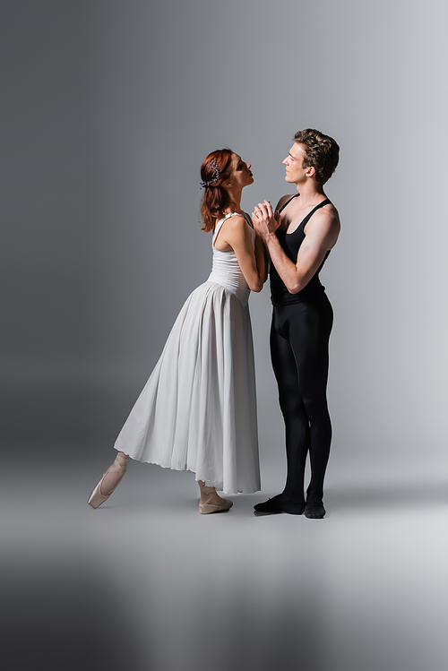 full length of graceful woman in white dress holding hands in ballet dancer on dark grey