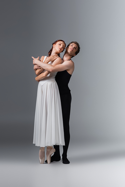 full length of ballet dancer hugging graceful ballerina in white dress on dark grey