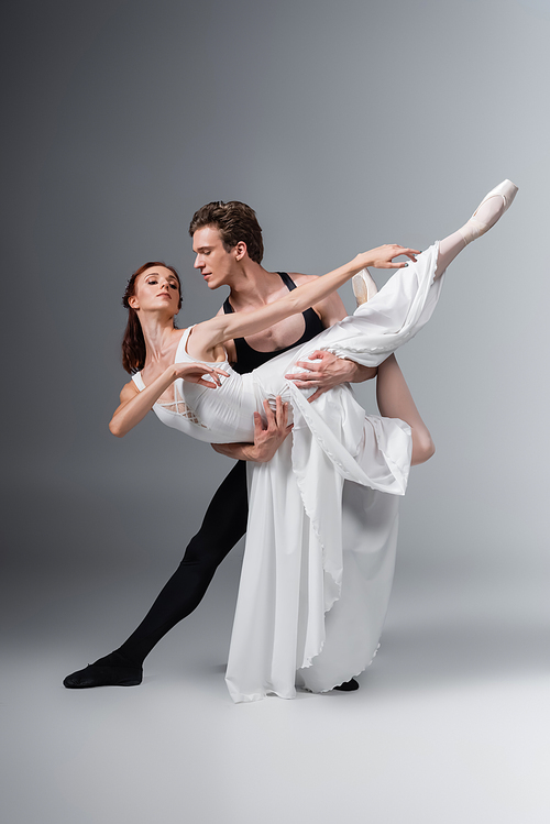 full length of man supporting graceful ballerina in white dress while dancing on dark grey