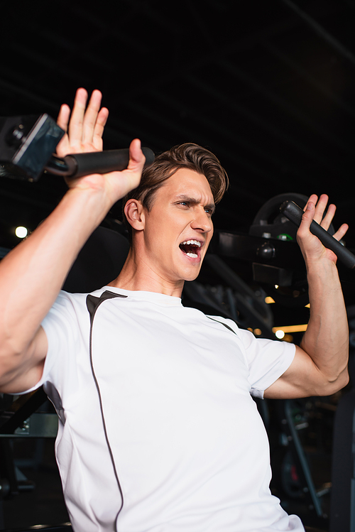 sportive man screaming while training on arm extension exercising machine in sports center