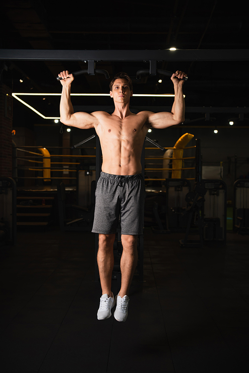 shirtless muscular man in shorts and sneakers working out on horizontal bar in gym