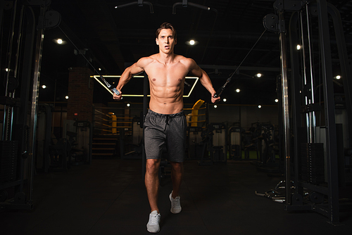 full length view of shirtless man in shorts training on pull rope exercising machine in gym