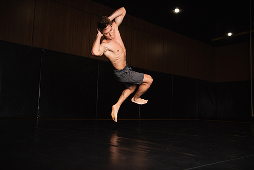 shirtless and barefoot sportsman in shorts jumping with hands behind head in gym