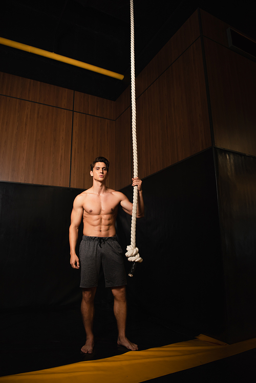 full length view of shirtless muscular man standing near gymnastic rope in gym