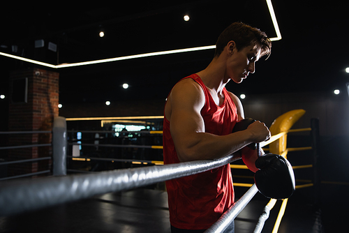 sportsman fastening boxing gloves near blurred ropes of ring in gym