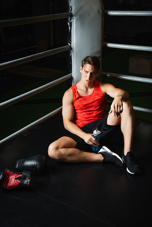 tired man in sportswear holding sports bottle while sitting on floor near boxing gloves