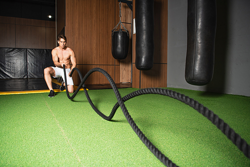 shirtless sportive man exercising with battle ropes in sports center