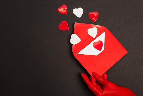 cropped view of person in red glove holding envelope with love letter and paper cut hearts on black