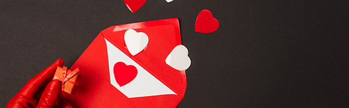cropped view of person in red glove holding tiny gift box near envelope and paper cut hearts isolated on black, banner