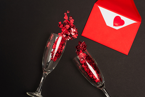 top view of champagne glasses with red confetti hearts near envelope on black