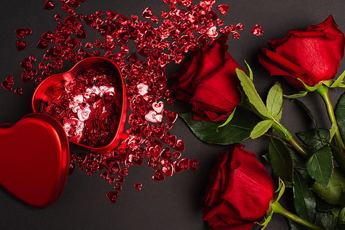 top view of red roses near metallic heart-shaped box and shiny confetti on black