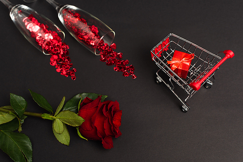 top view of red rose near glasses with confetti and tiny present in shopping cart on black
