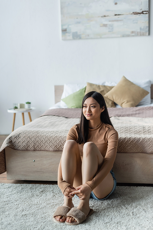 smiling asian woman sitting on floor near bed and looking away