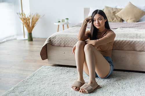 young asian woman looking away while sitting on floor in bedroom