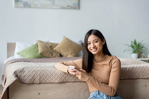 joyful asian woman smiling at camera while using smartphone in bedroom
