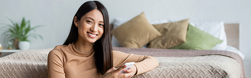 cheerful asian woman  while using smartphone in bedroom, banner
