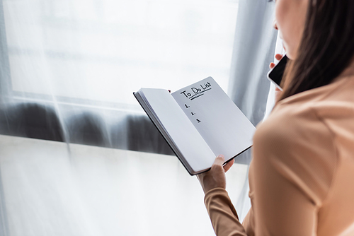 cropped view of woman holding notebook with to do list lettering while talking on smartphone