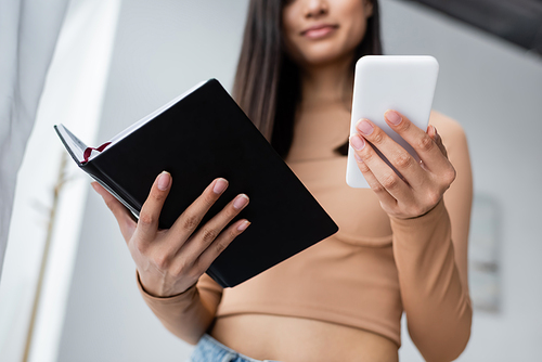 cropped view of blurred woman holding smartphone and notebook while working at home