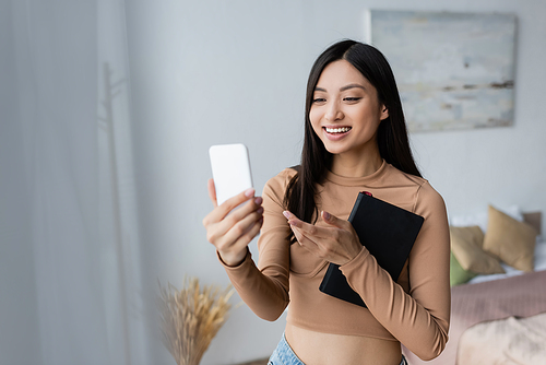 smiling asian woman with copy book pointing with finger during video call on cellphone