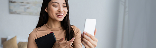 happy asian woman with notebook pointing with finger during video chat on smartphone, banner