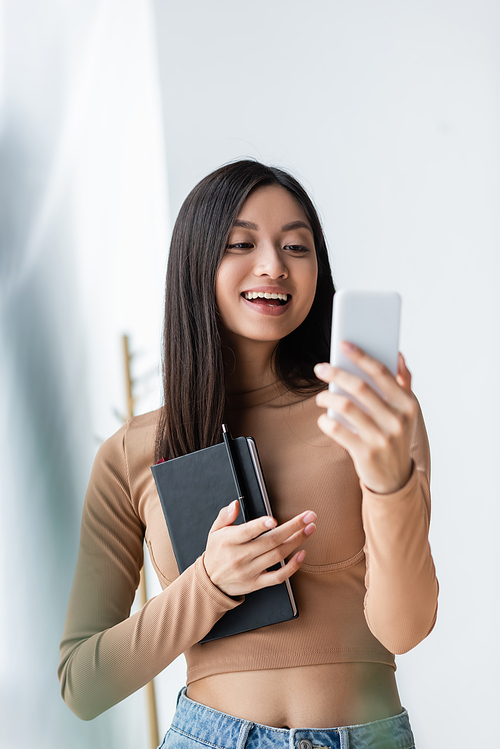cheerful asian woman holding notebook and pointing with finger during video call on smartphone
