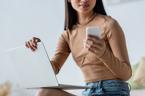 cropped view of young woman using smartphone near laptop at home