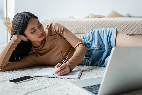 young asian woman writing in notebook while lying on floor near blurred laptop