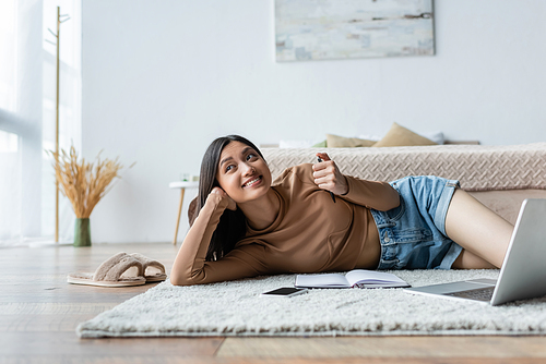 smiling asian woman lying on carpet in bedroom near gadgets and notebook