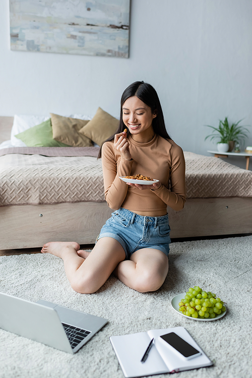 happy asian freelancer eating almonds near laptop and snacks on floor in bedroom
