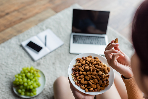 cropped view of woman eating almonds near blurred gadgets and fresh grape on floor