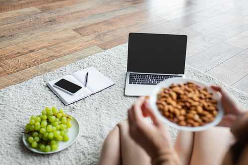 partial view of blurred freelancer holding bowl with almonds near gadgets and fresh grape on floor