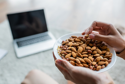 partial view of woman with bowl of almonds near blurred laptop with blank screen