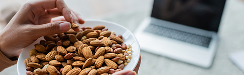 partial view of woman with bowl of almonds near blurred laptop with blank screen, banner