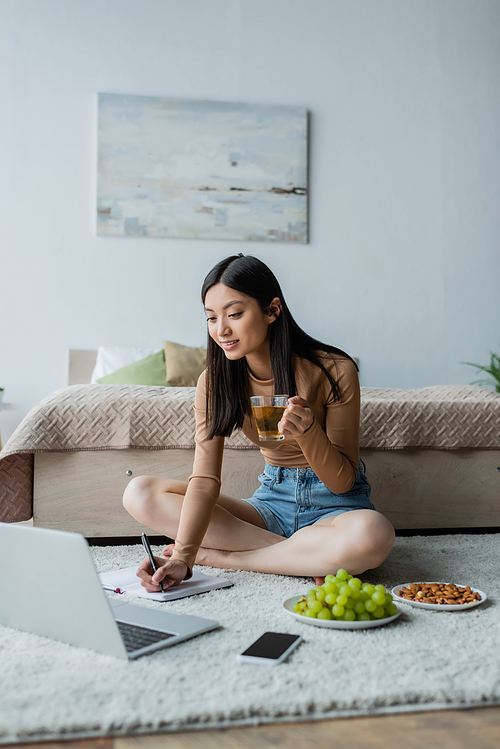 asian woman with cup of tea writing in notebook near gadgets, almonds and grape on floor