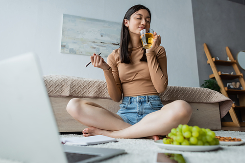 pleased woman enjoying flavor of tea near blurred laptop and grape of floor
