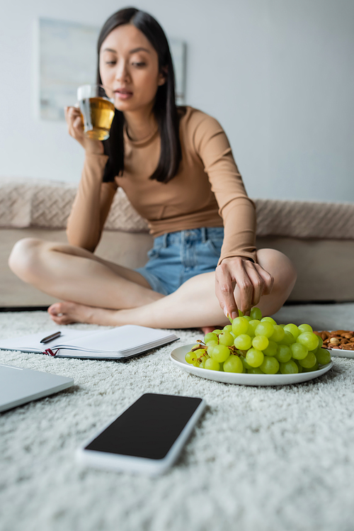 blurred asian woman taking grape while drinking tea on floor near notebook and smartphone