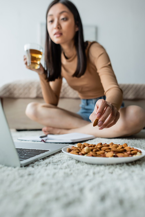 blurred asian woman drinking tea and eating almonds while looking at laptop on floor