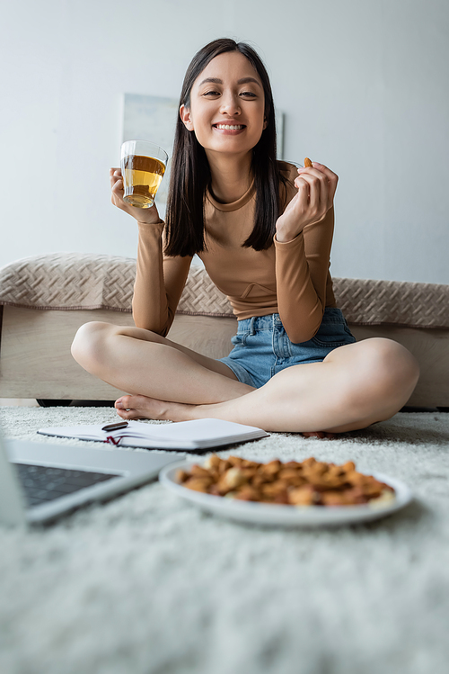 happy asian woman eating almonds and drinking tea on floor near blurred laptop
