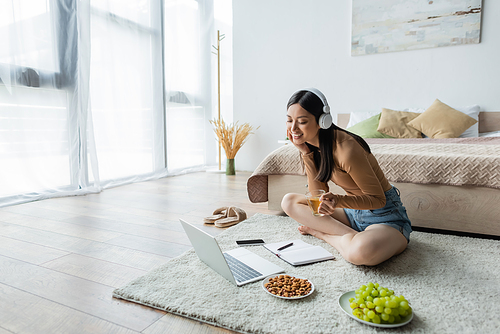 happy asian woman in headphones sitting on floor with cup of tea near gadgets and snacks